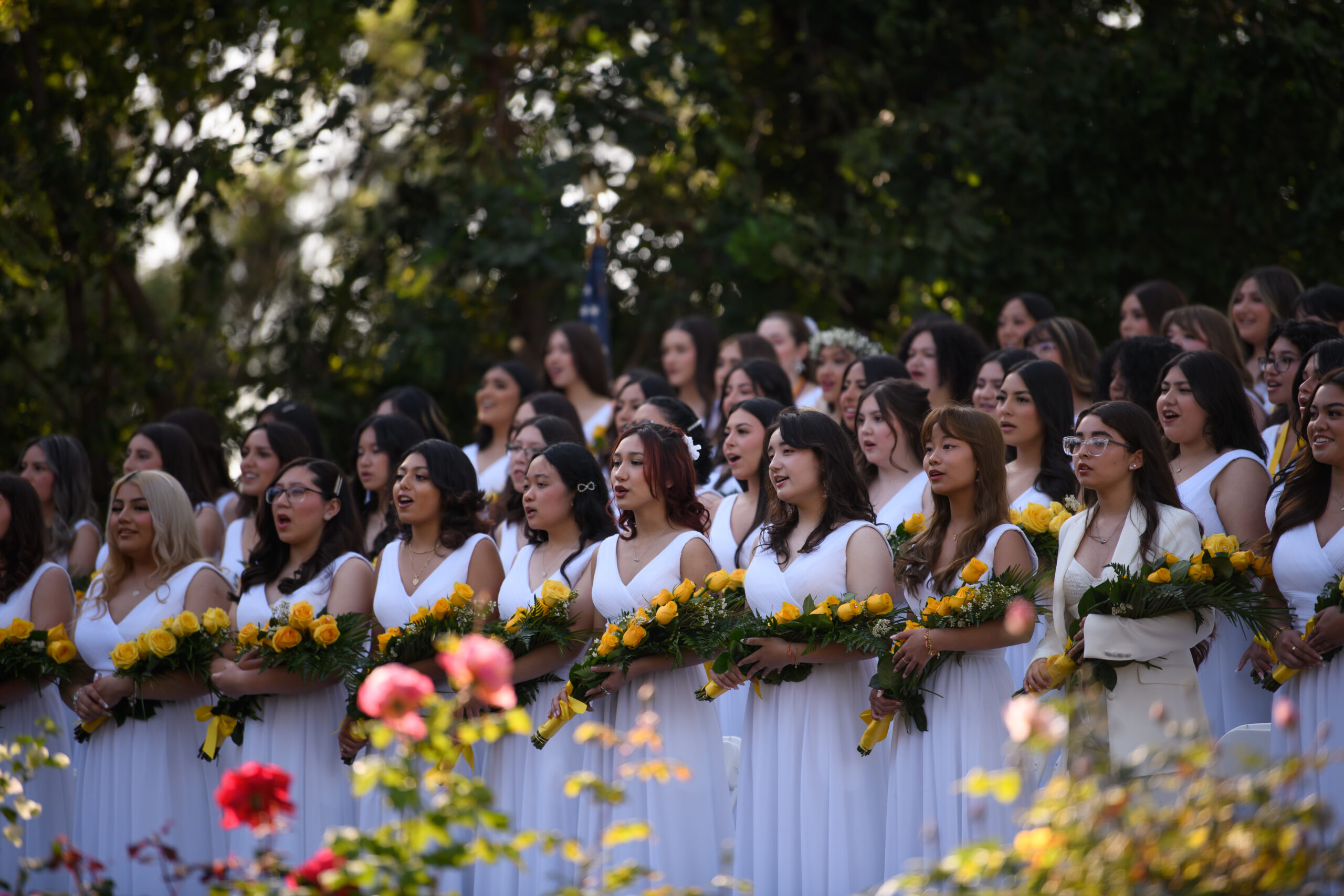 Ramona Convent graduates wearing white dresses and holding yellow flowers, lined up in rows at the graduation ceremony.